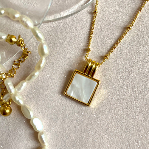 Square Mother of pearl pendant set in yellow gold on a 18ct yellow gold plated bobble chain 20 inch in total. Part of a glass and see pearl necklace in part of the picture on a cream background